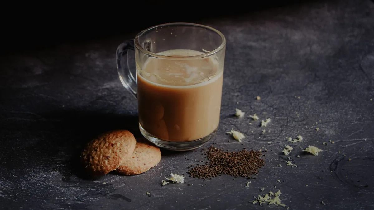 A glass mug of milky tea beside two round biscuits and scattered tea leaves on a dark stone surface