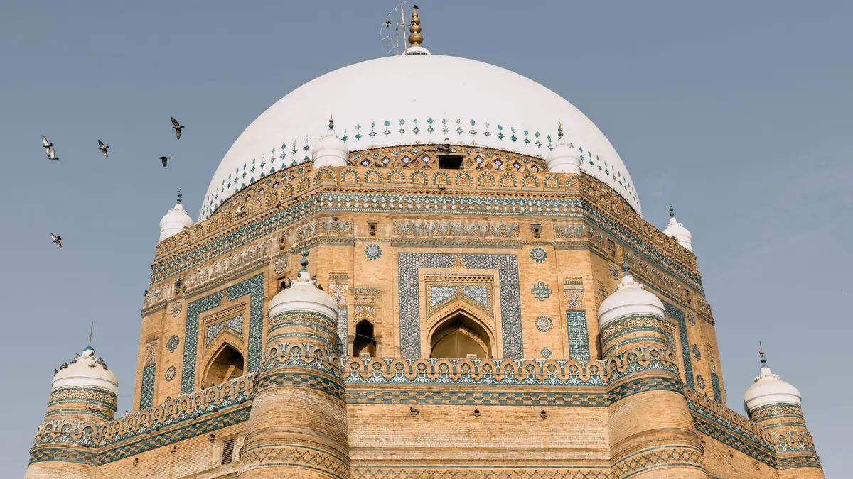 Red brick octagonal tomb with blue tilework and a white dome under a clear sky, pigeons flying overhead