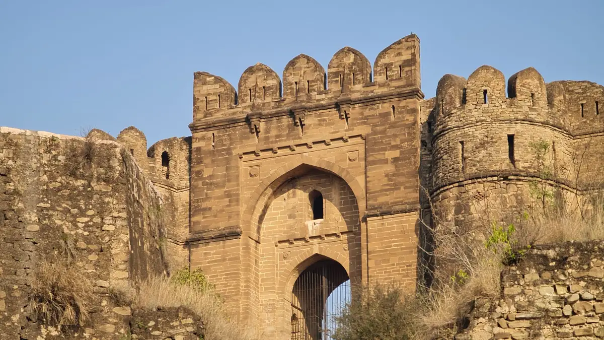 Sandstone gateway of Rohtas Fort with crenellated towers, an arched entrance and dry grass on the walls under a blue sky