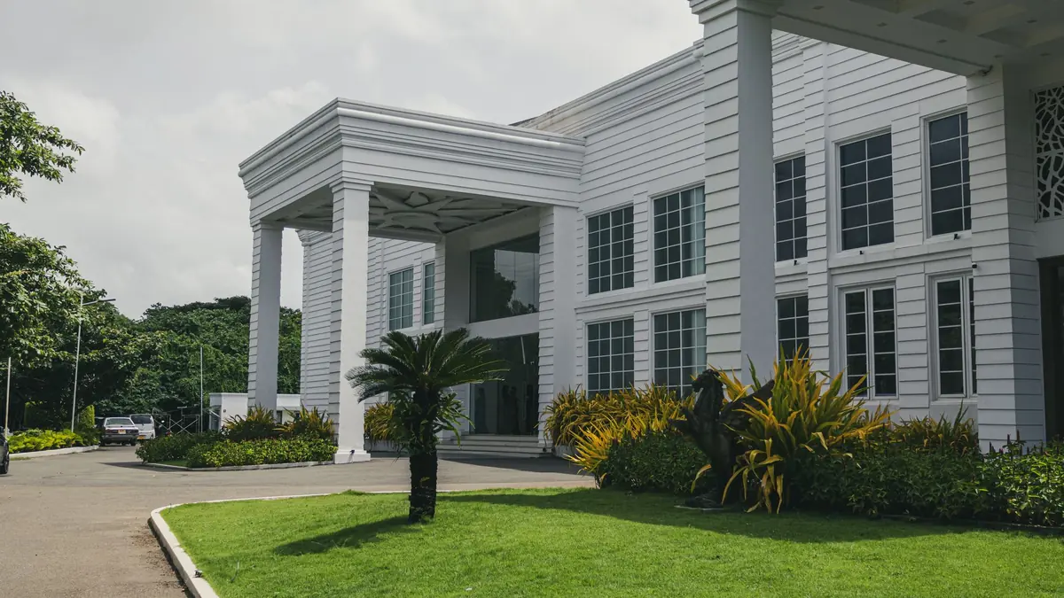 White hotel building with a pillared entrance porch, curved driveway, palm and landscaped lawn under a cloudy sky