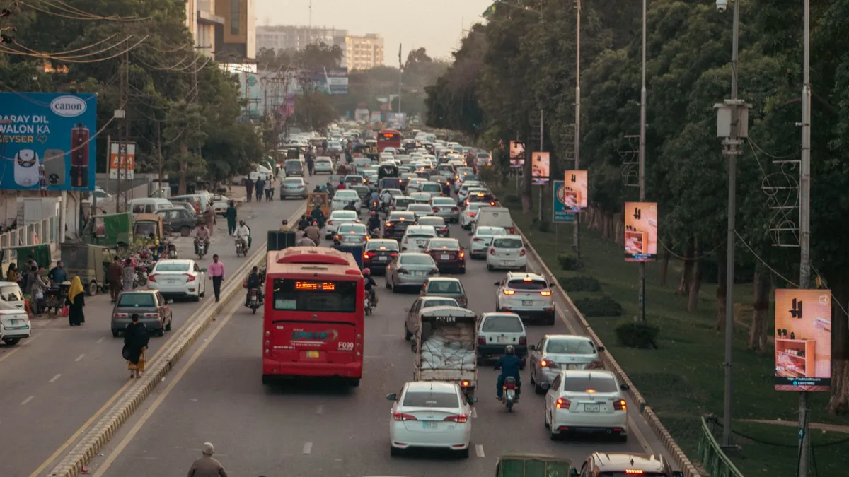 Busy tree-lined arterial road with cars, a red bus and motorbikes in slow traffic under hazy daylight