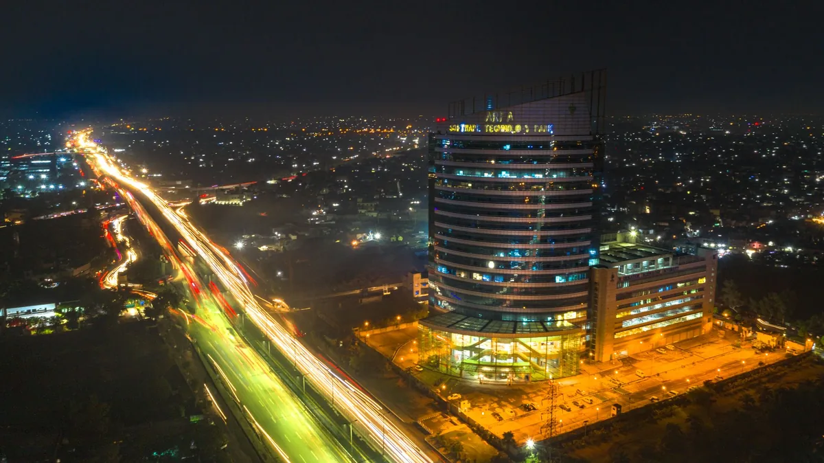 Aerial night view of a lit office tower beside a busy Lahore road with long streaks of vehicle light trails