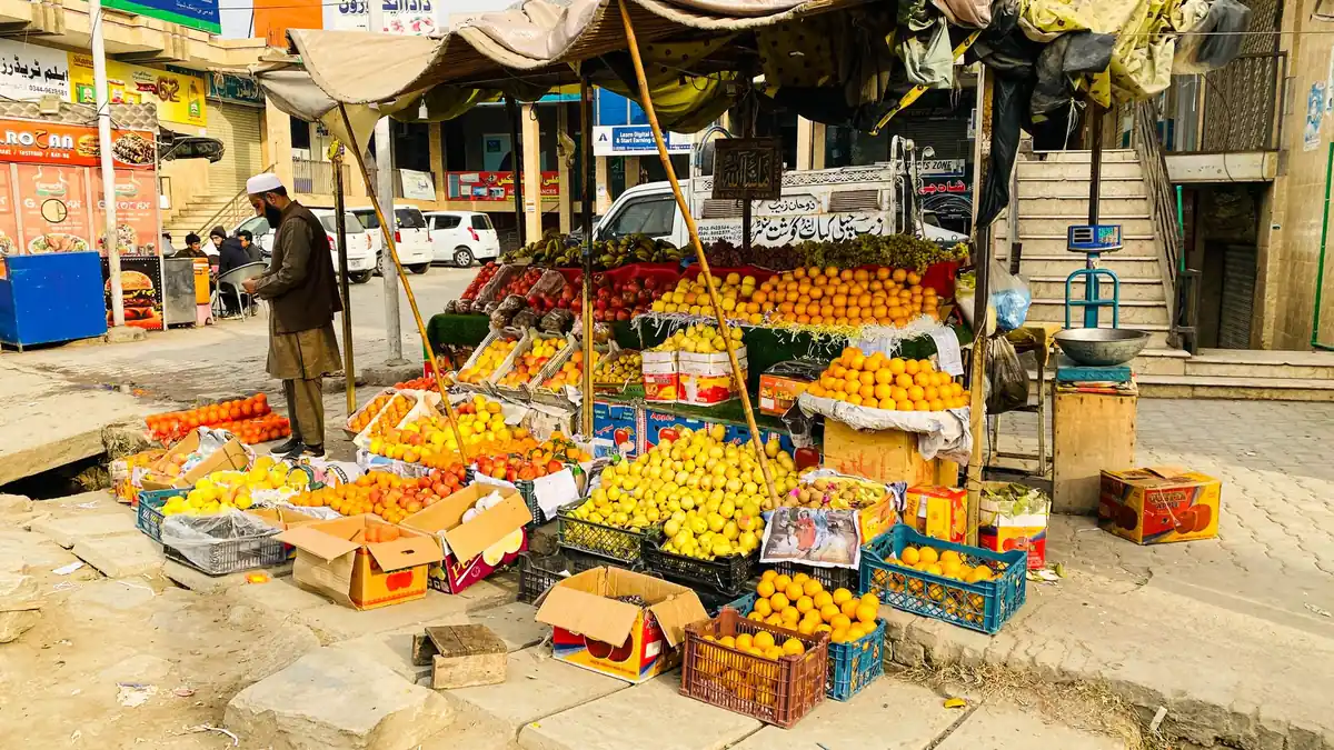 Roadside fruit stall piled with oranges, apples and bananas in crates under a canopy on a Pakistani bazaar street