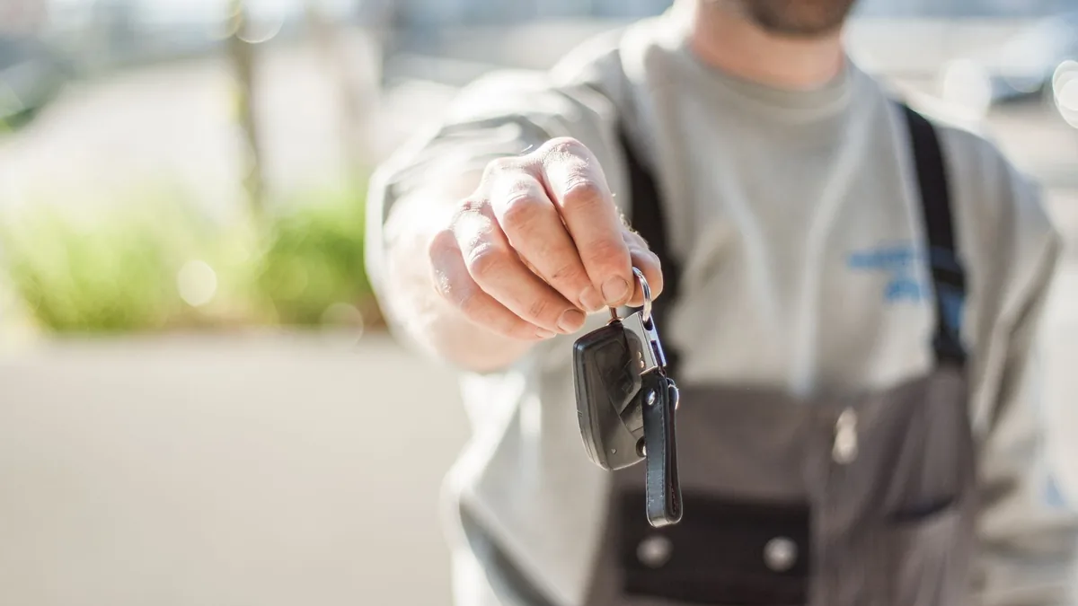 Man in grey work overalls holds out a single car key towards the camera, face cropped out of frame, outdoors