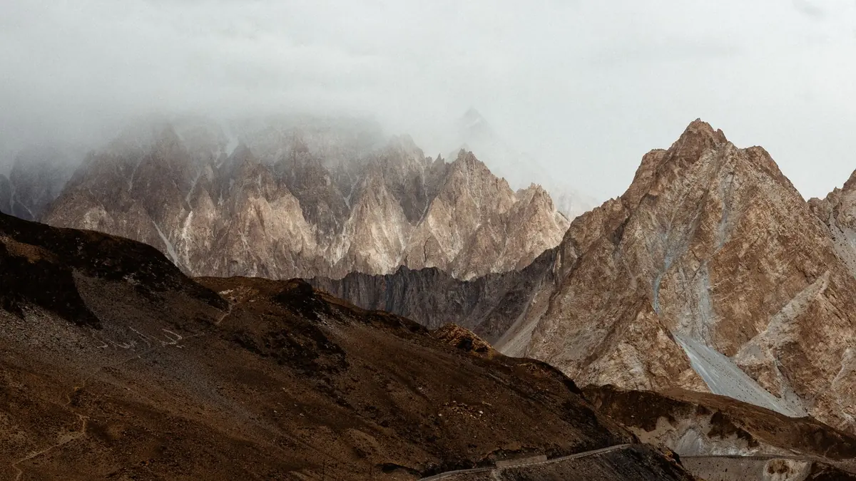 Jagged rocky peaks under low cloud above a narrow road winding along a bare brown mountainside