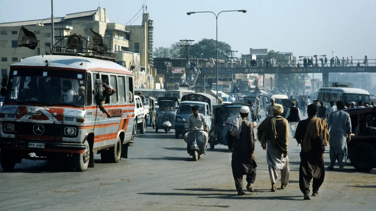 Busy Pakistani city street with a passenger minibus, cars, motorbikes and pedestrians under a footbridge