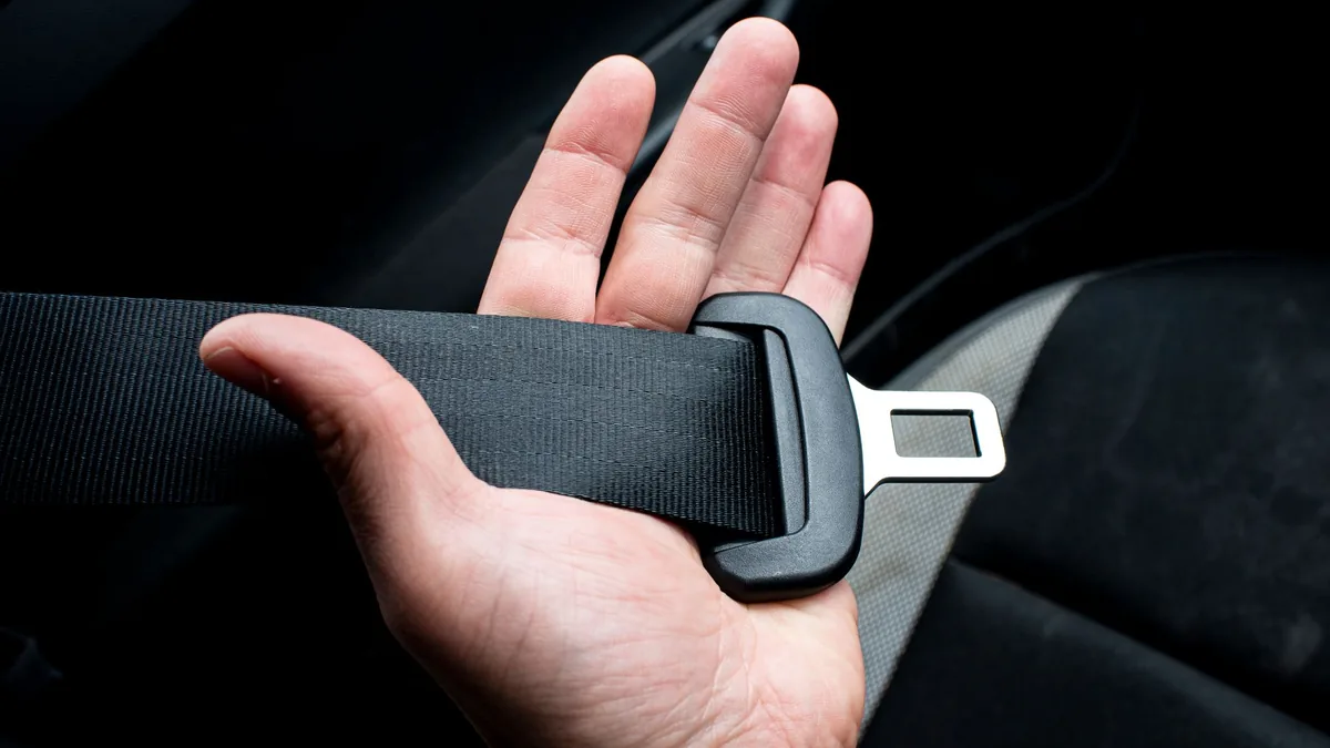Close-up of an open hand holding a car seat belt tongue against a dark fabric car seat