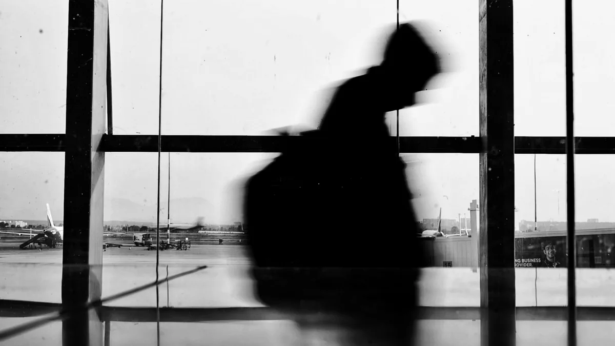 Black-and-white silhouette of a traveller with a backpack walking past airport terminal windows, parked aircraft outside