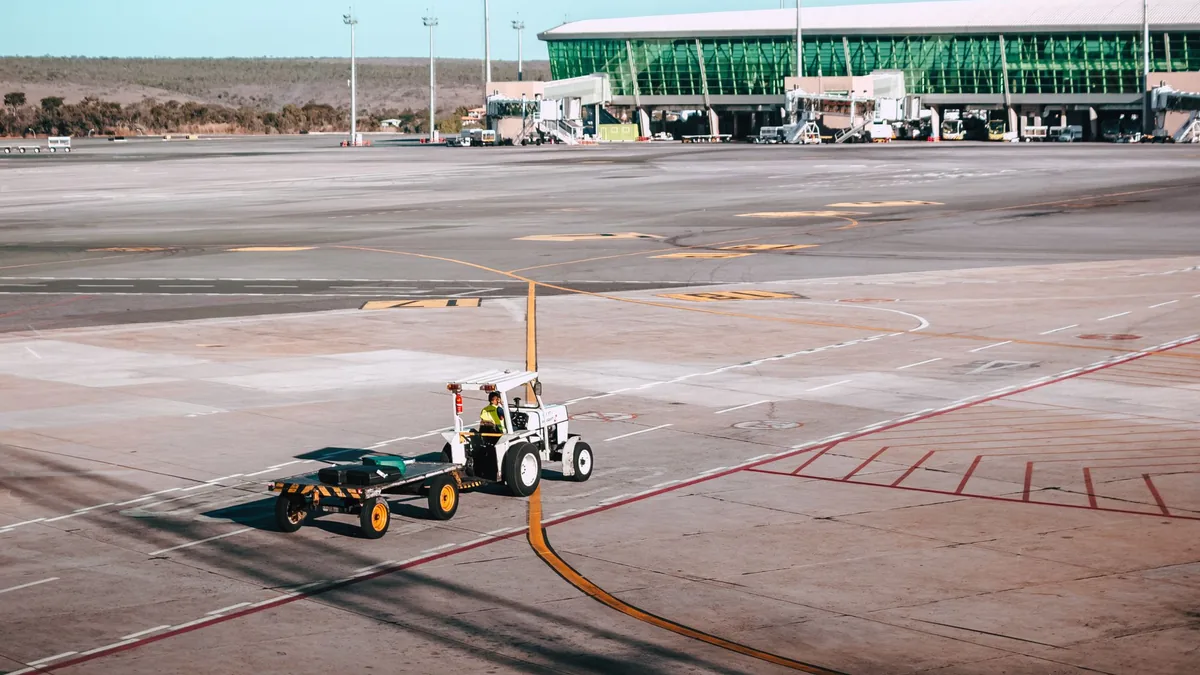 Baggage tractor towing an empty luggage cart across an airport apron beside a glass-fronted terminal on a clear day