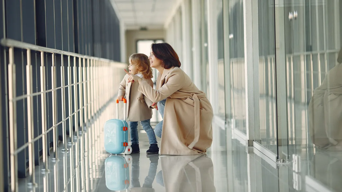 Mother and young child with a small blue suitcase in a bright, glass-walled terminal corridor