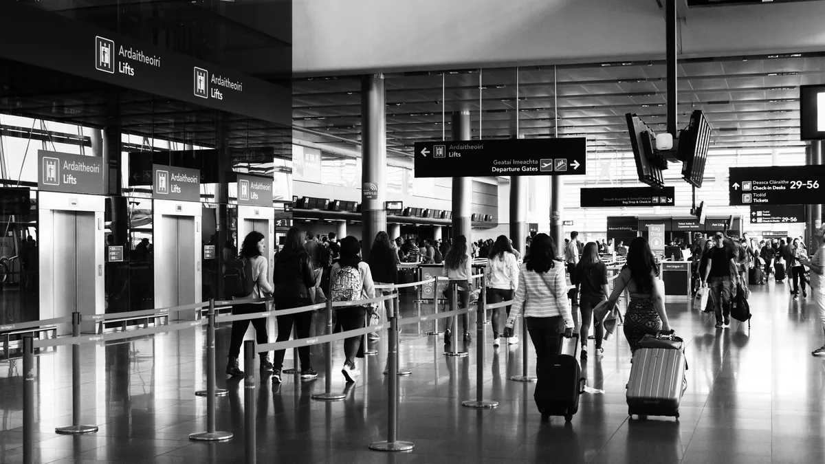 Black and white view of travellers pulling suitcases through a busy airport terminal hall past queue barriers