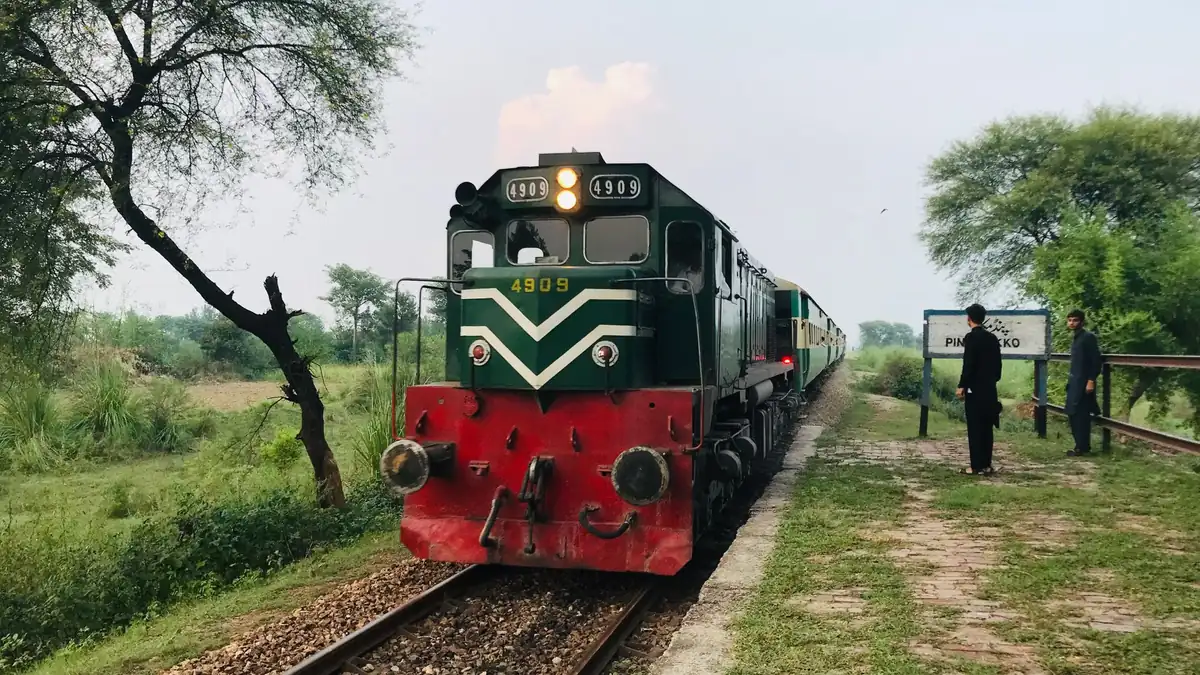 Green and red diesel locomotive hauling a passenger train along a tree-lined rural track beside a small platform