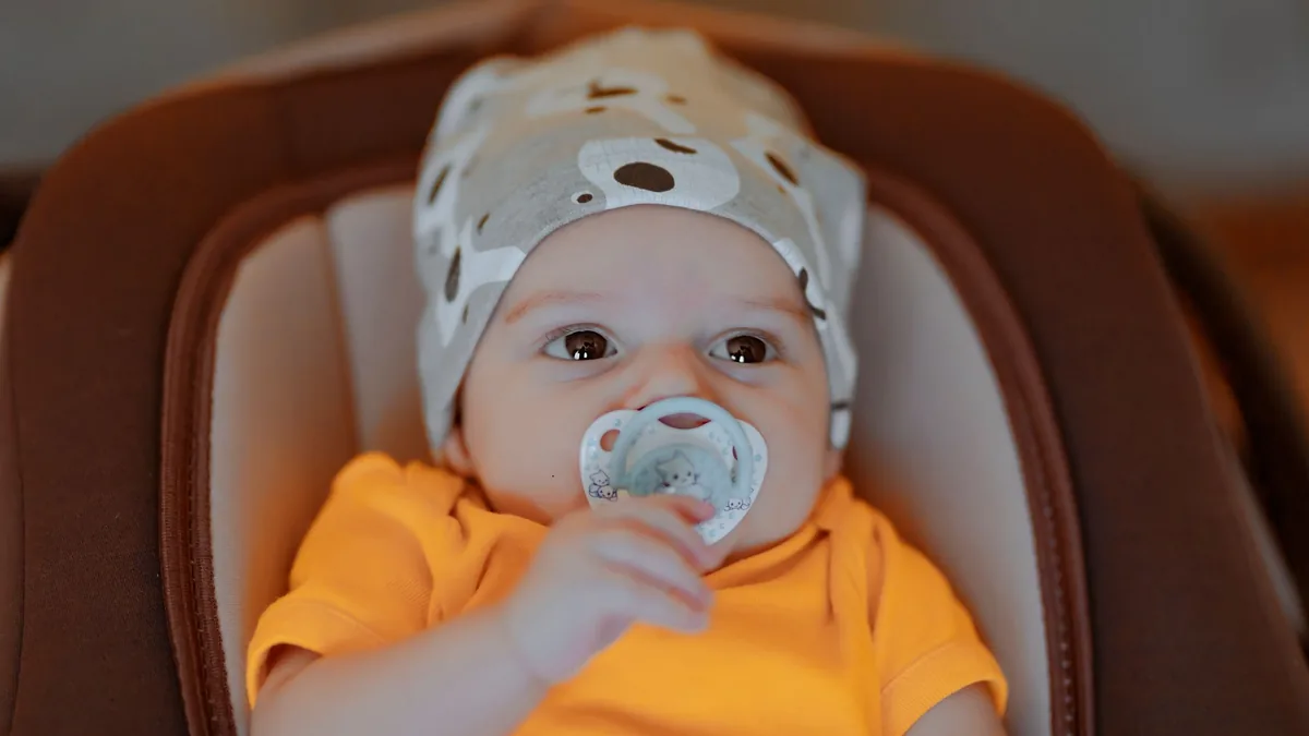 Baby in a knitted hat and orange top sitting in a padded child seat with a dummy in its mouth