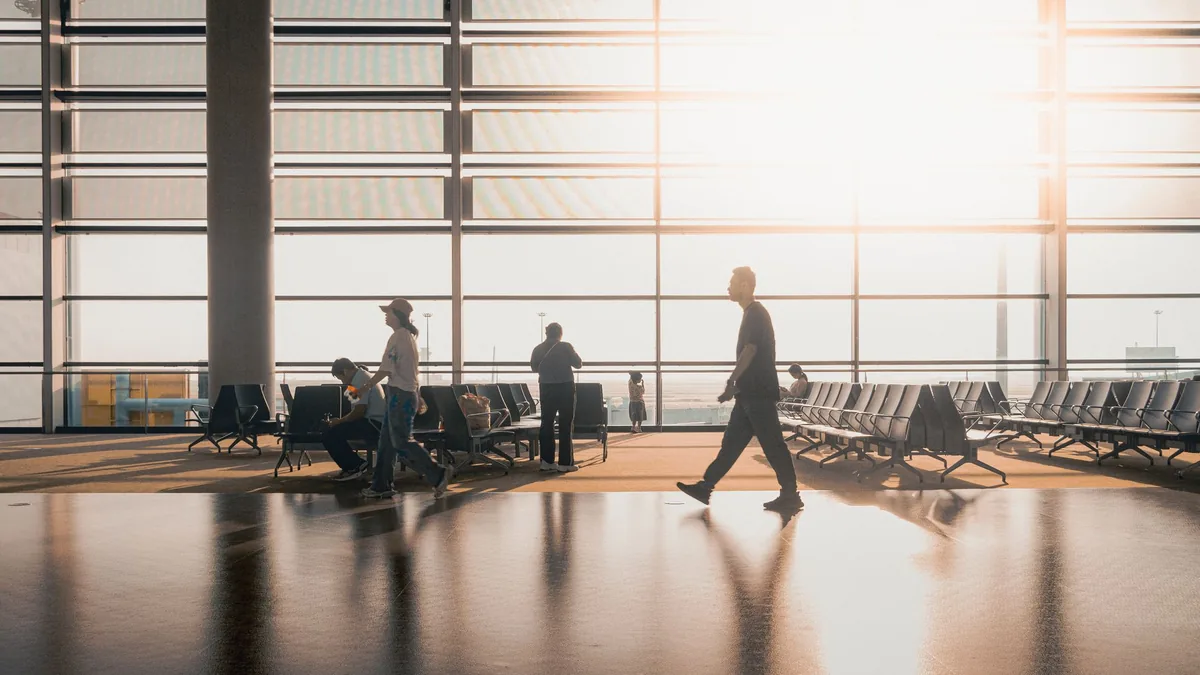 Silhouetted travellers walking and seated in a sunlit airport terminal hall with tall glass windows