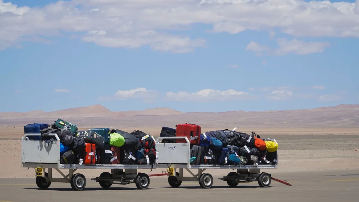 Two airport baggage carts piled high with colourful suitcases and holdalls under a clear blue sky