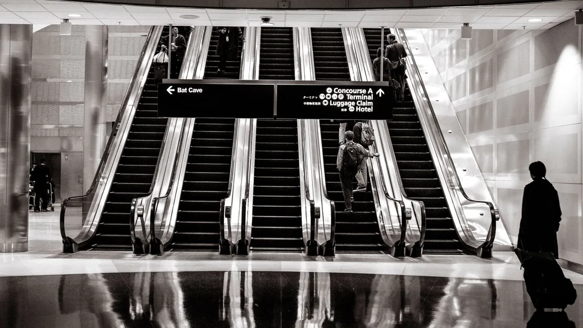 Black and white view of travellers on a bank of escalators in a modern airport terminal beneath overhead direction signs