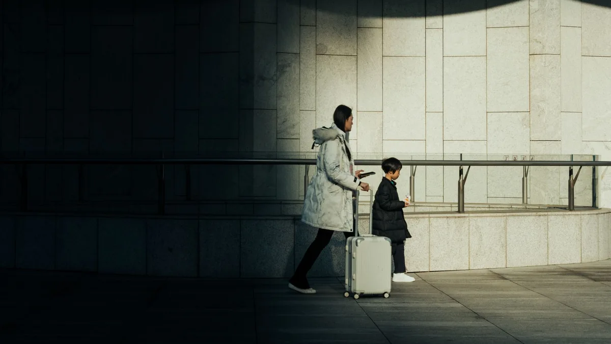 An adult and a small child with a wheeled suitcase walk through a dim, empty concrete hall lit by a patch of daylight