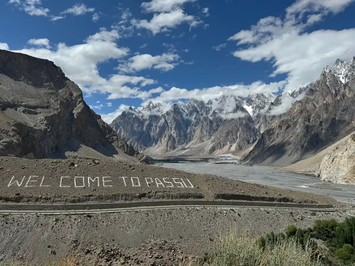 A welcome sign on the Karakoram Highway with the Passu Cones behind it